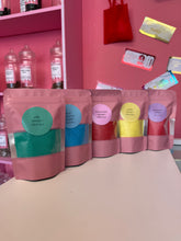 Load image into Gallery viewer, Five stand-up pouches of sherbet crystals in different colors (green, blue, red, yellow) lined up on a light pink surface against a pink background.
